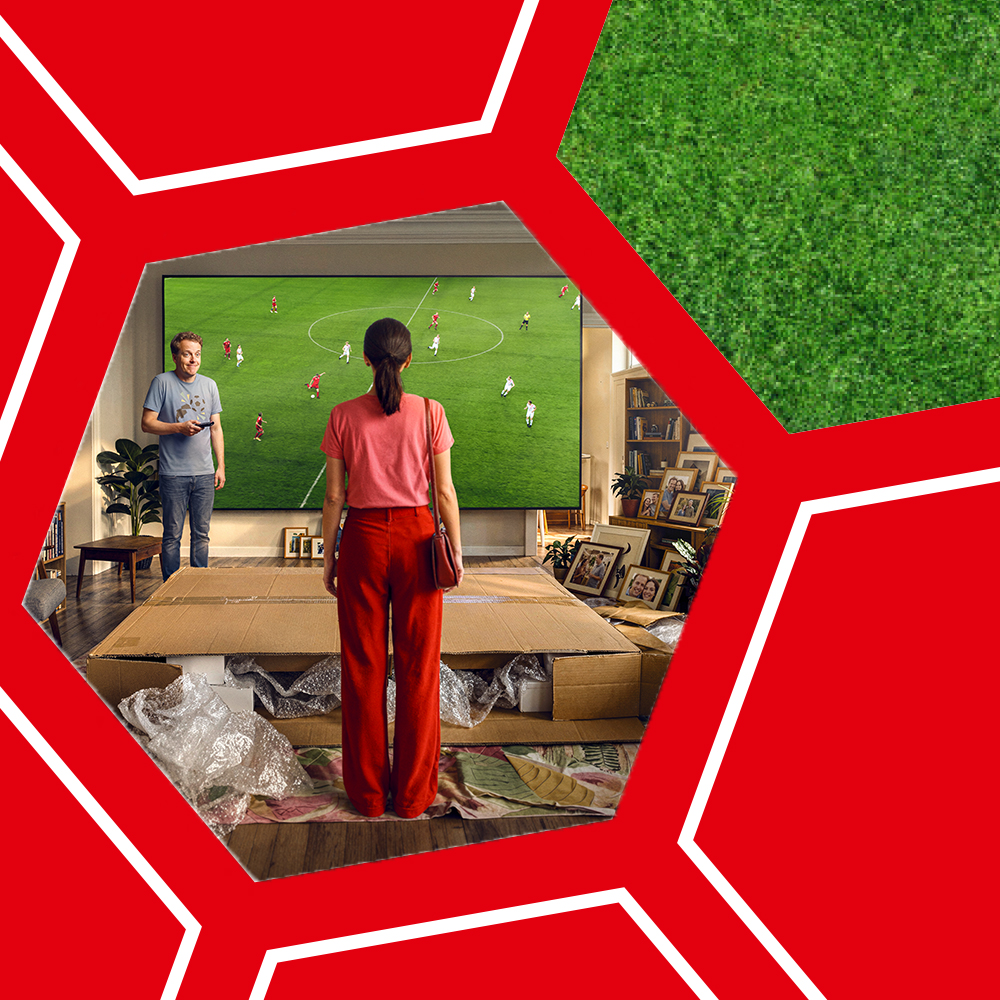 Zwei Personen stehen vor einem großen Fernseher, auf dem ein Fußballspiel gezeigt wird, umgeben von roten Fünfecken und grünem Rasenmuster